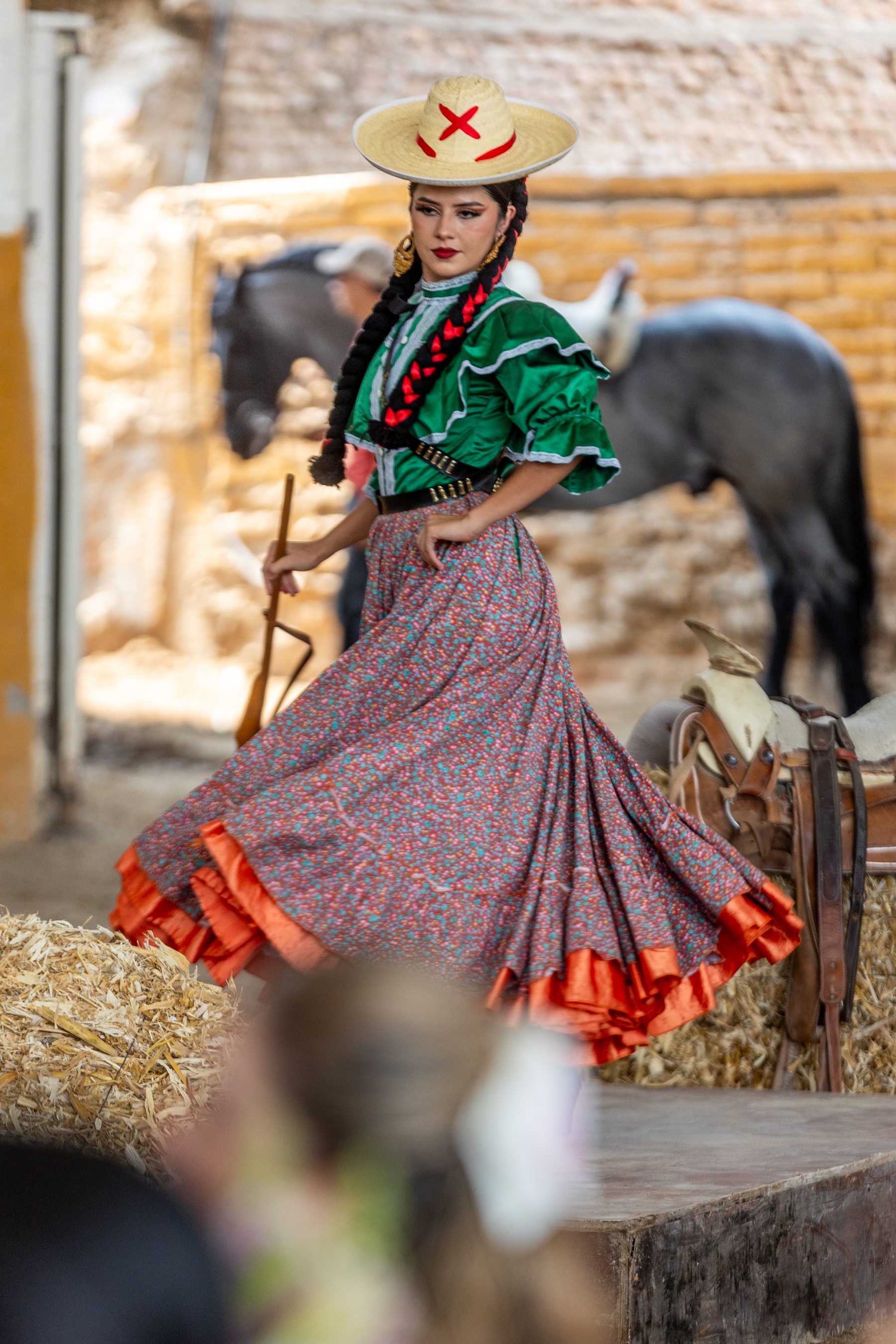 Escaramuza con traje verde y falda floreada posando en el rancho con caballo al fondo